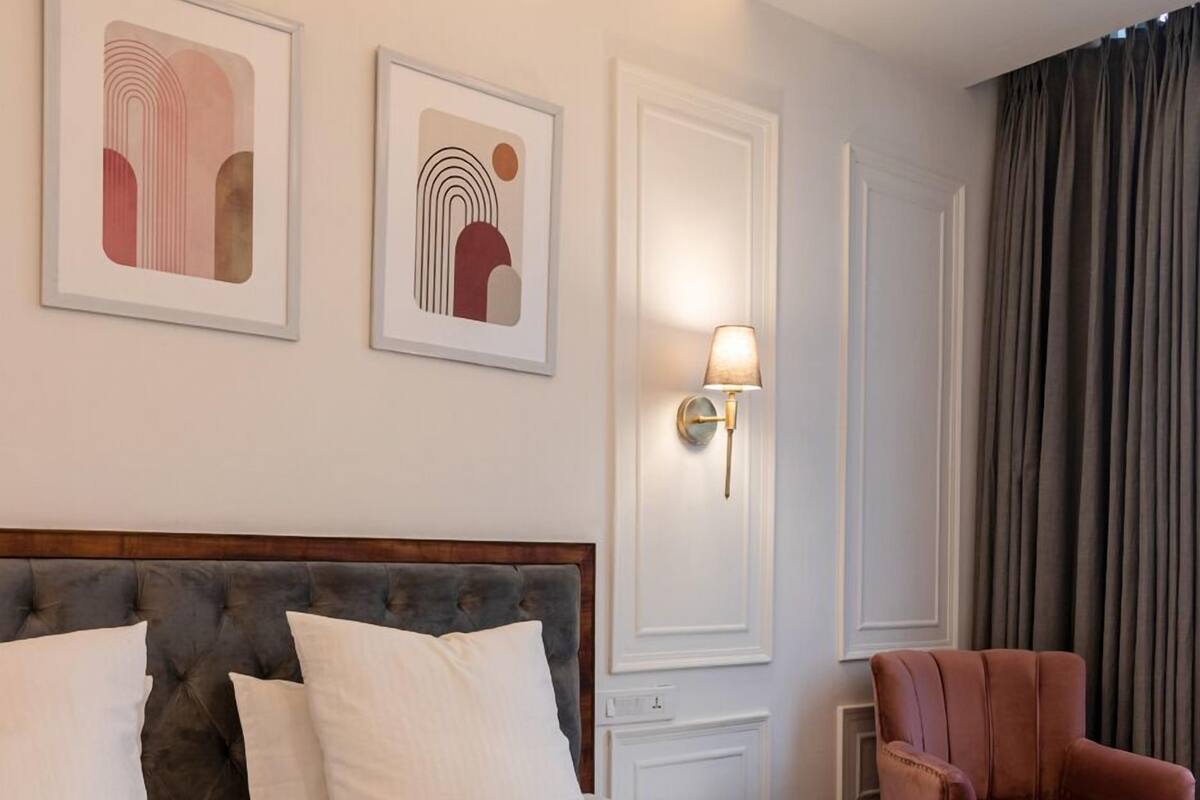 A hotel room with a large bed, a headboard, two wall-mounted artworks, a bedside lamp, a chair, and a window with curtains.