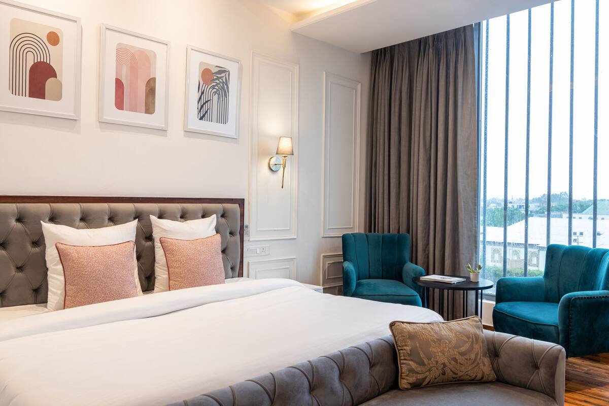 A hotel room with a large bed, two armchairs, a small table, and a view of the city through large windows.