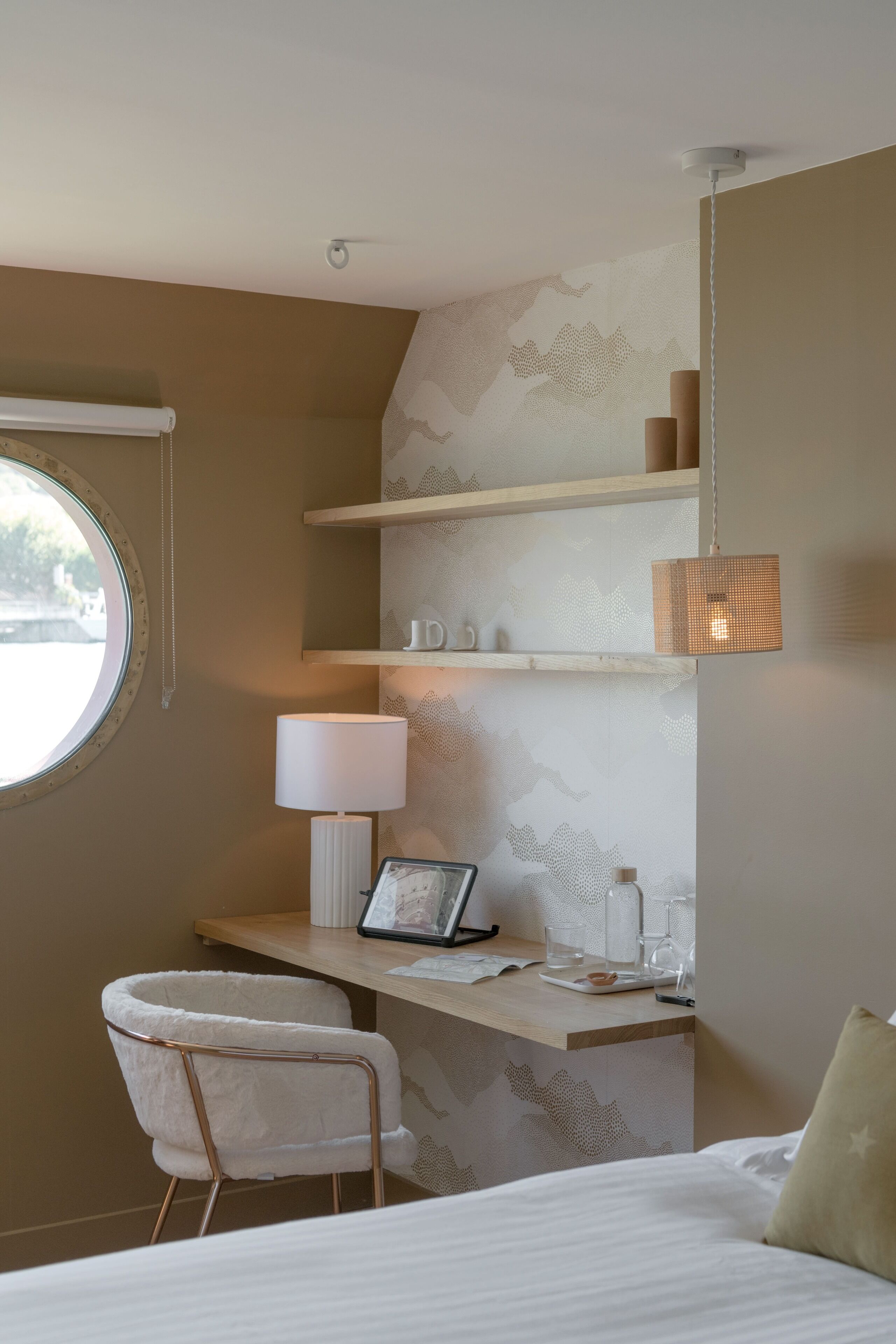 A hotel room with a bed, a desk with a chair, a lamp, and shelves with decorative items.