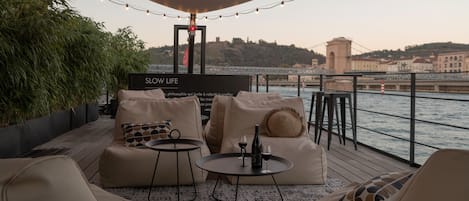 Un espace extérieur aménagé avec des canapés et des tables, une enseigne indiquant « SLOW LIFE », et une vue sur une rivière et des bâtiments en arrière-plan.