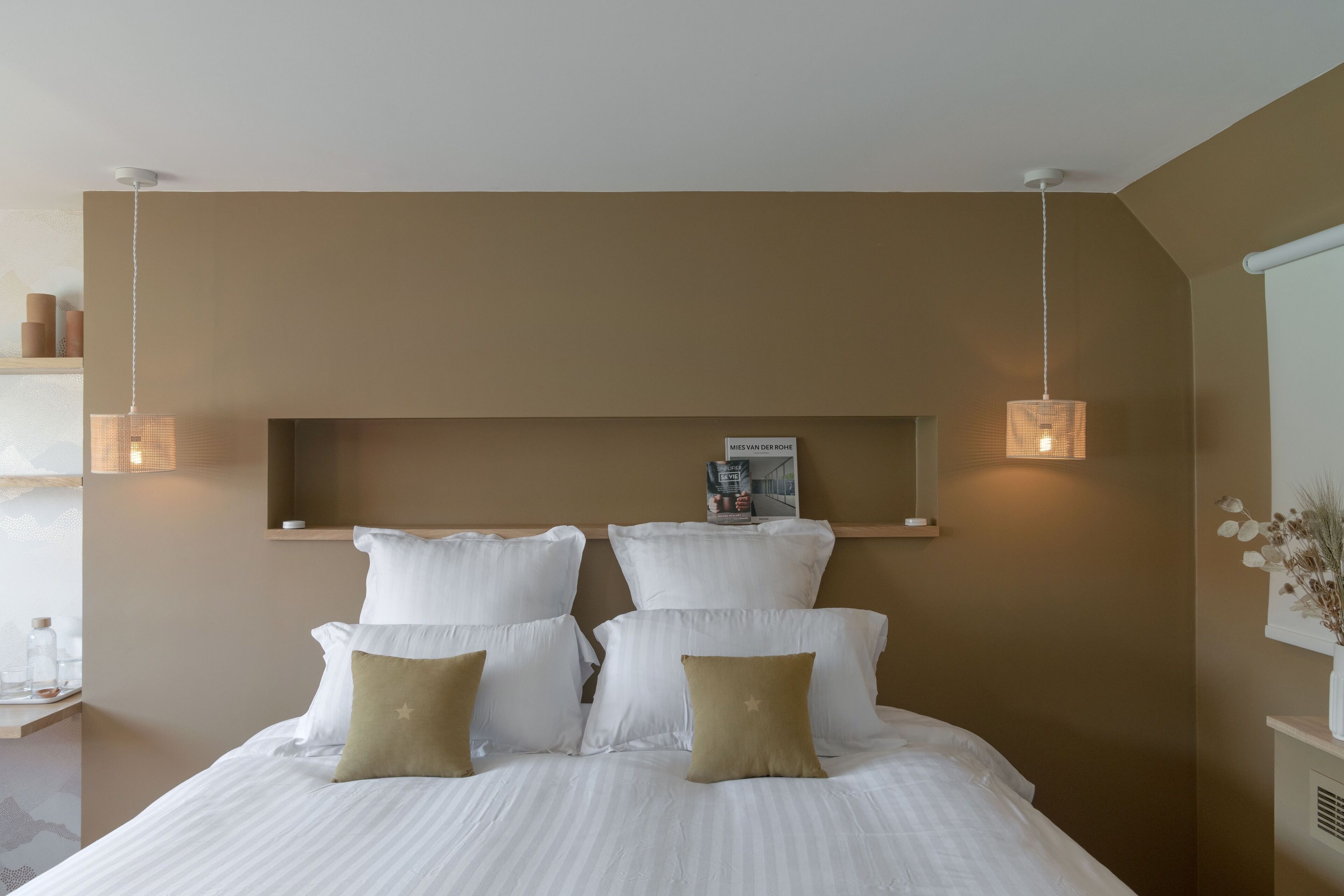 A neatly made bed with white and gold pillows, a magazine on the headboard, and two pendant lights.