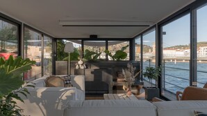 Een modern, ruim balkon met uitzicht op het water en gebouwen, ingericht met witte banken, een haardvuur en diverse planten.