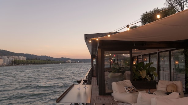 Een terras met zitplaatsen en een tafel, met uitzicht op een waterpartij en gebouwen op de achtergrond.