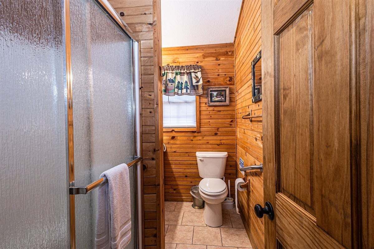 A compact wooden bathroom with a toilet, a towel rack, and a window with a curtain.