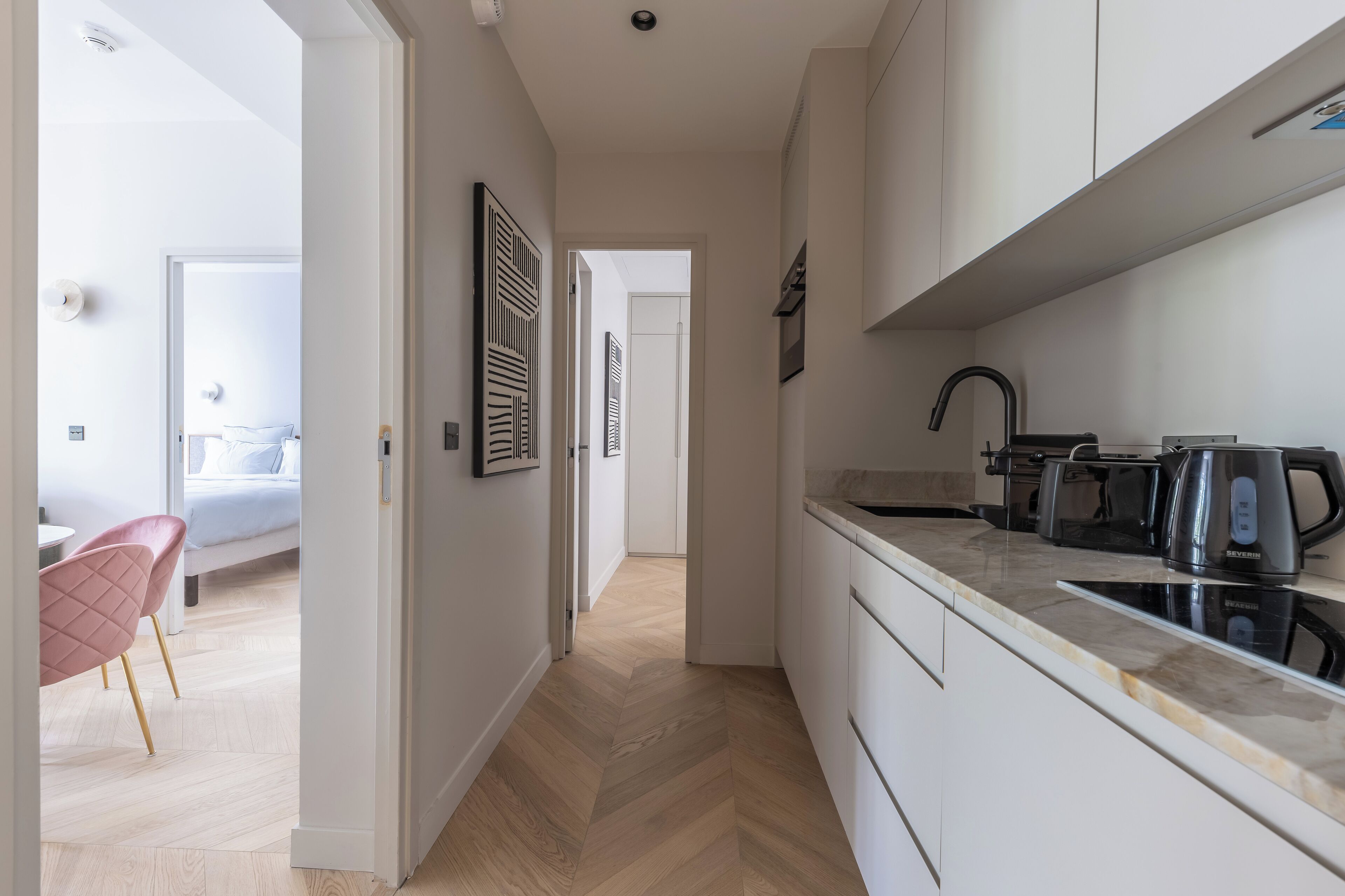 A modern kitchen with white cabinets, a black faucet, and a kettle. A wooden herringbone floor leads to a bedroom with a bed and a pink chair.
