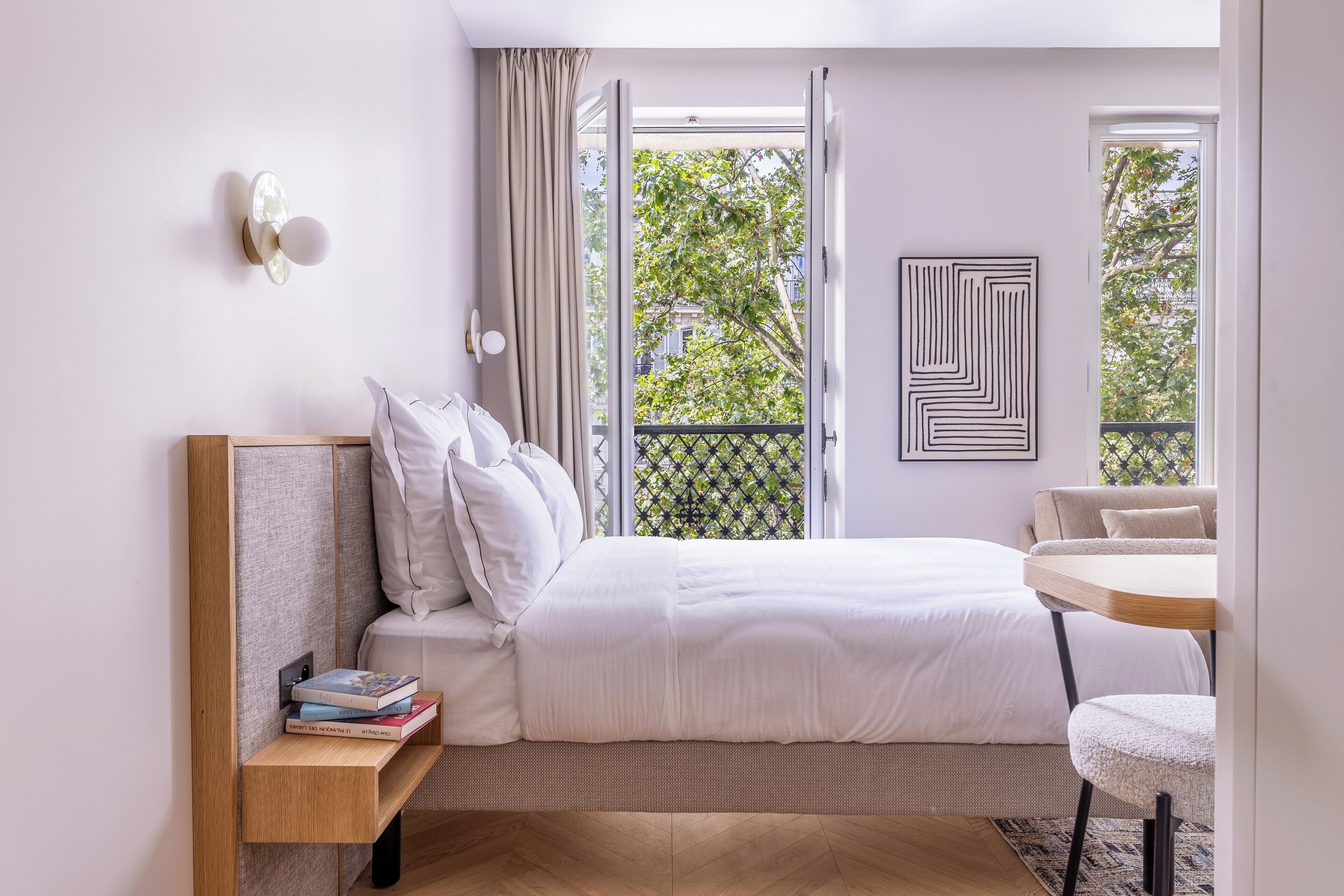 A modern bedroom with a large bed, a bedside table with books, a small round table, and a view of greenery through the window.