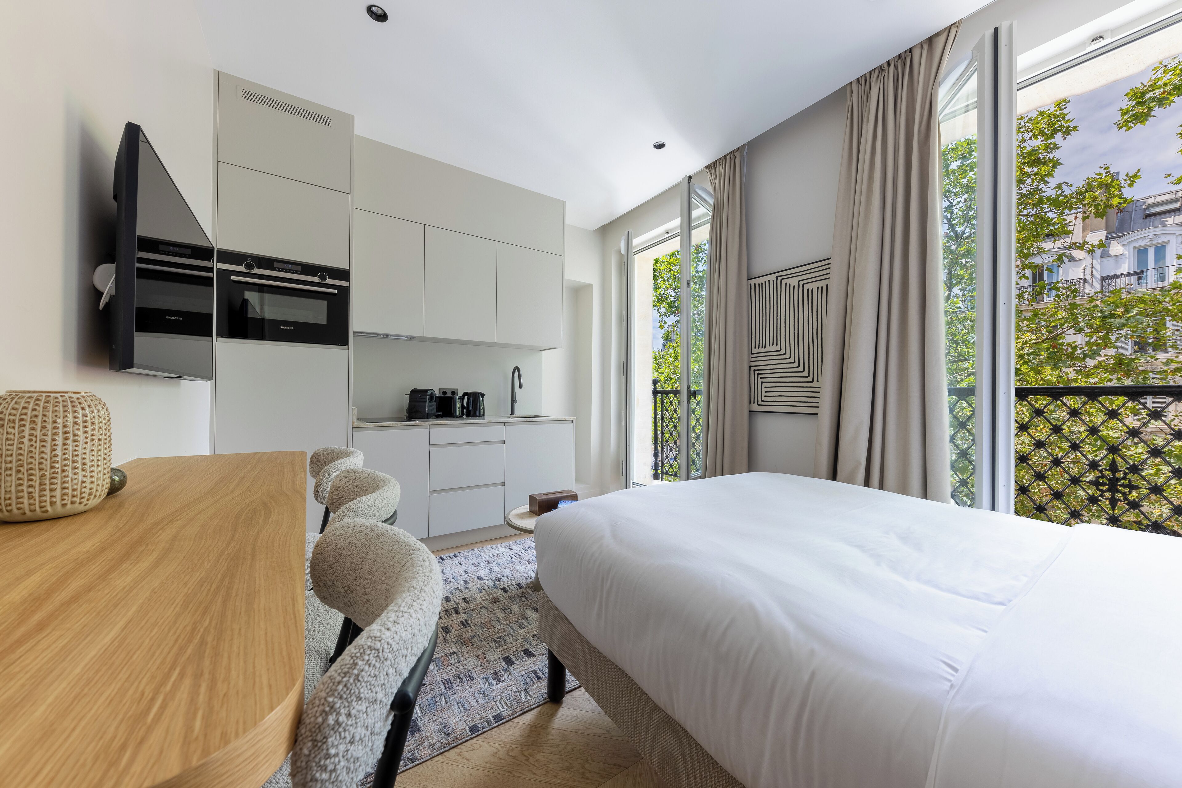 A modern hotel room with a large bed, a desk, a kitchenette, and a balcony with a view of the city.