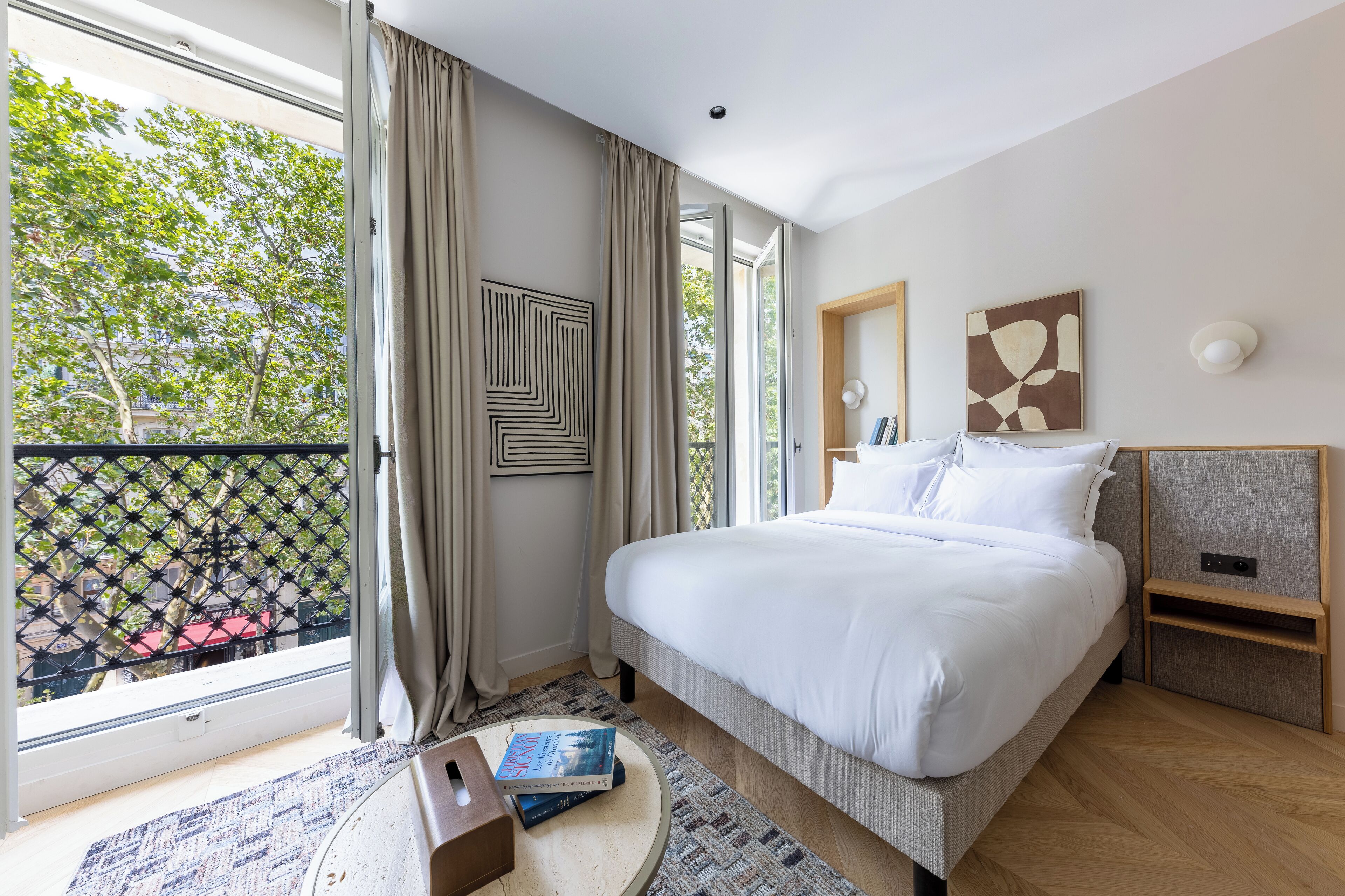 A modern hotel room with a large bed, a small bedside table, a window with curtains, and a view of trees and buildings outside.
