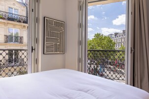 A hotel room with a large bed, a view of the city through a balcony, and a geometric artwork on the wall.