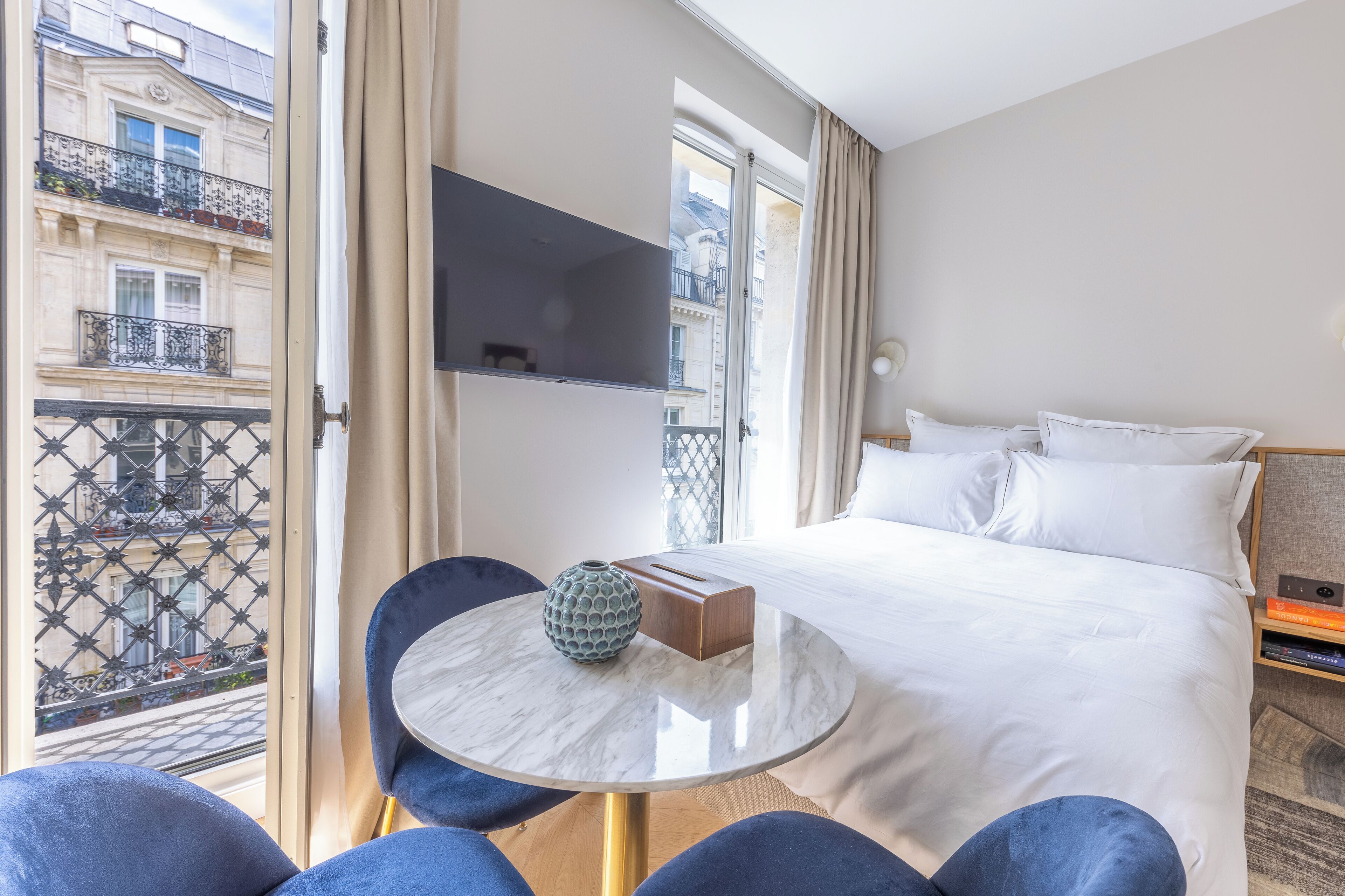A hotel room with a bed, a TV mounted on the wall, a round table with a vase, and a view of the city through a balcony.