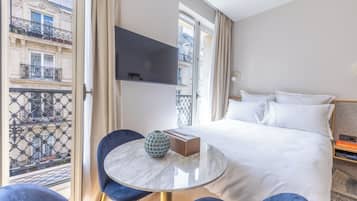 A hotel room with a bed, a TV mounted on the wall, a round table with a vase, and a view of the city through a balcony.
