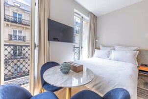 A hotel room with a bed, a TV mounted on the wall, a round table with a vase, and a view of the city through a balcony.