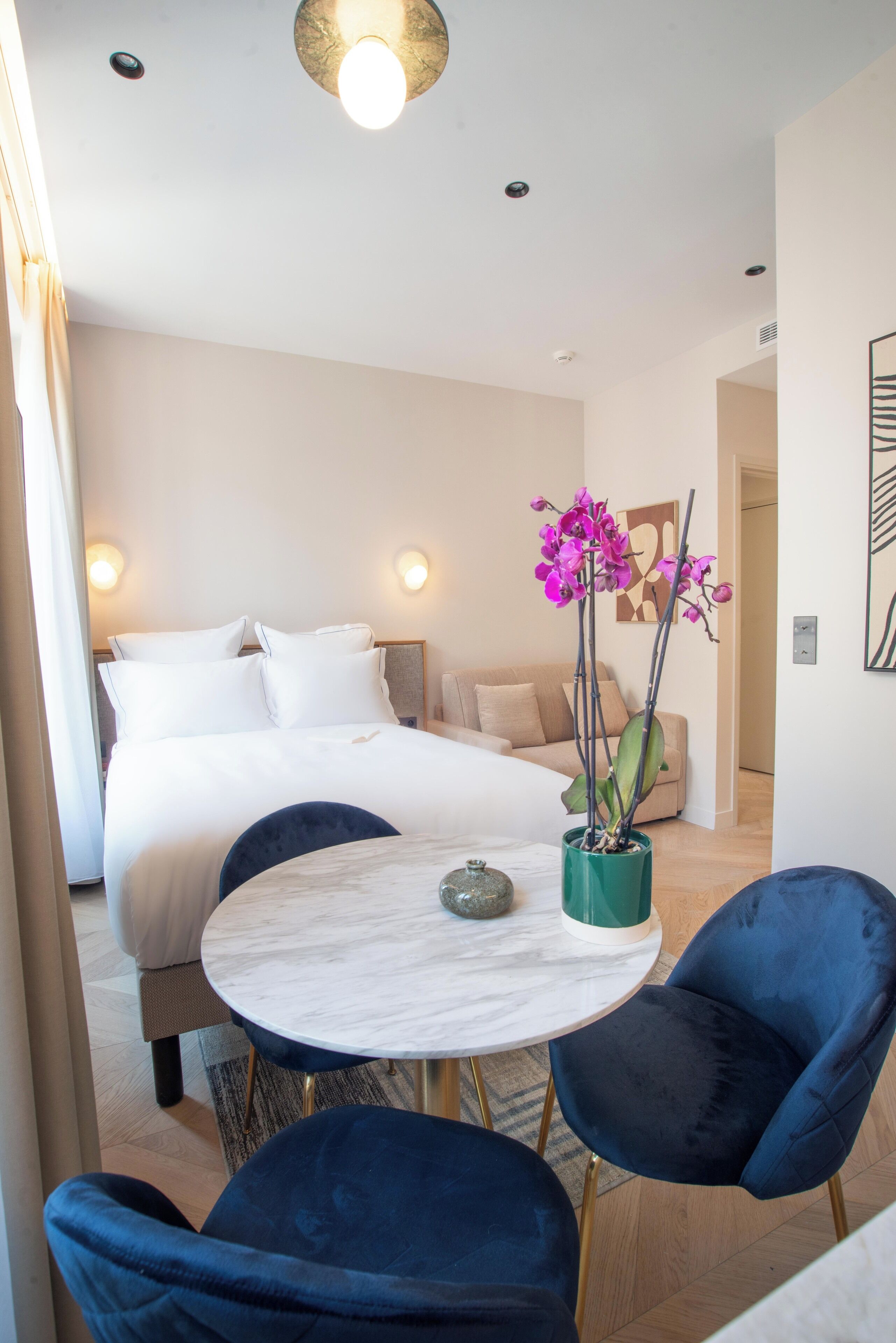 A modern hotel room with a bed, a round table with chairs, a potted orchid, and a painting on the wall.