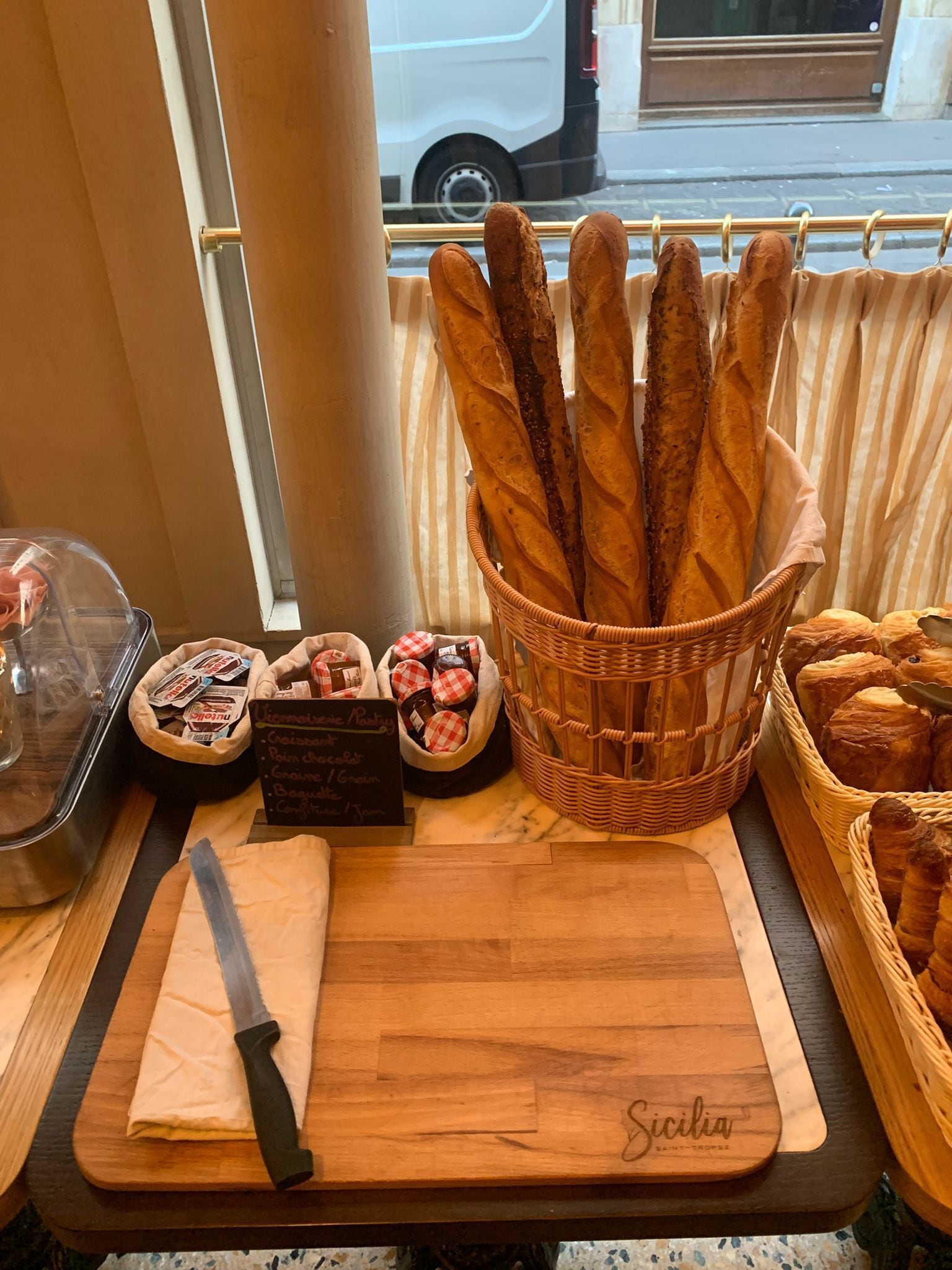 A wooden cutting board with a knife, a basket of baguettes, and a sign listing bakery items.