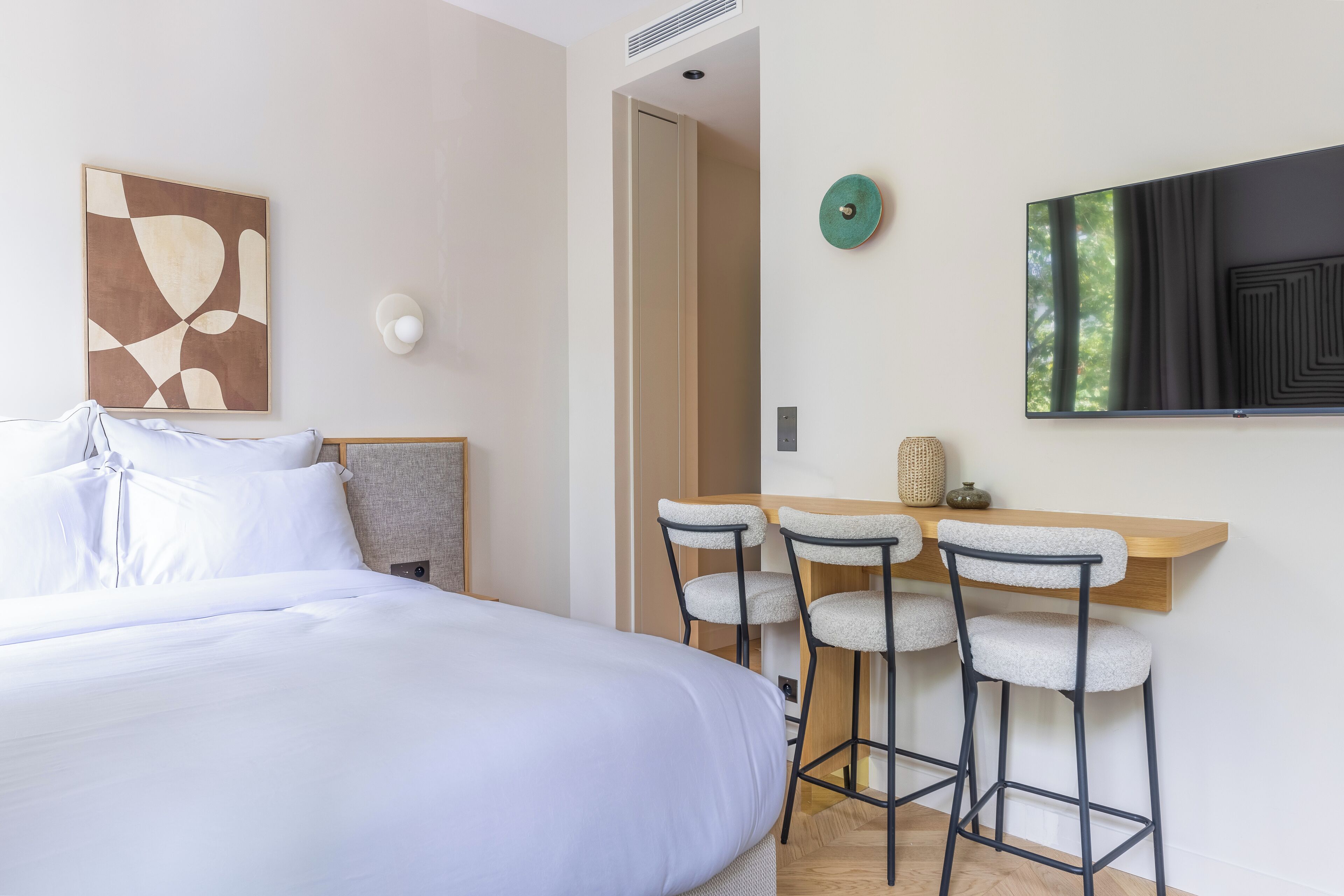 A modern hotel room with a large bed, a desk with bar stools, a flat-screen TV, and a wall-mounted artwork.