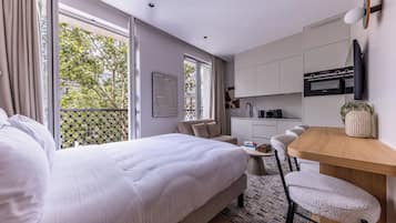 A modern hotel room with a large bed, a dining area with a wooden table and bar stools, a kitchenette, and a balcony with a view of trees and buildings.