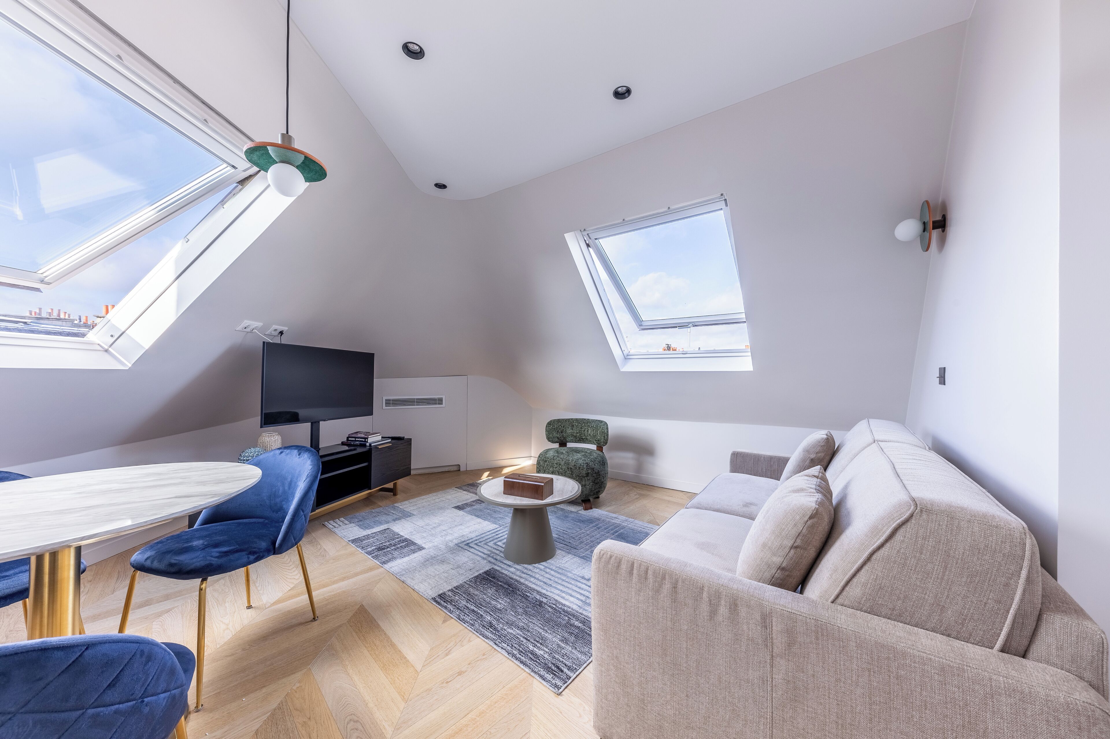 A modern living room with a high ceiling, skylights, a flat-screen TV, a dining table with chairs, a sofa, and a coffee table.