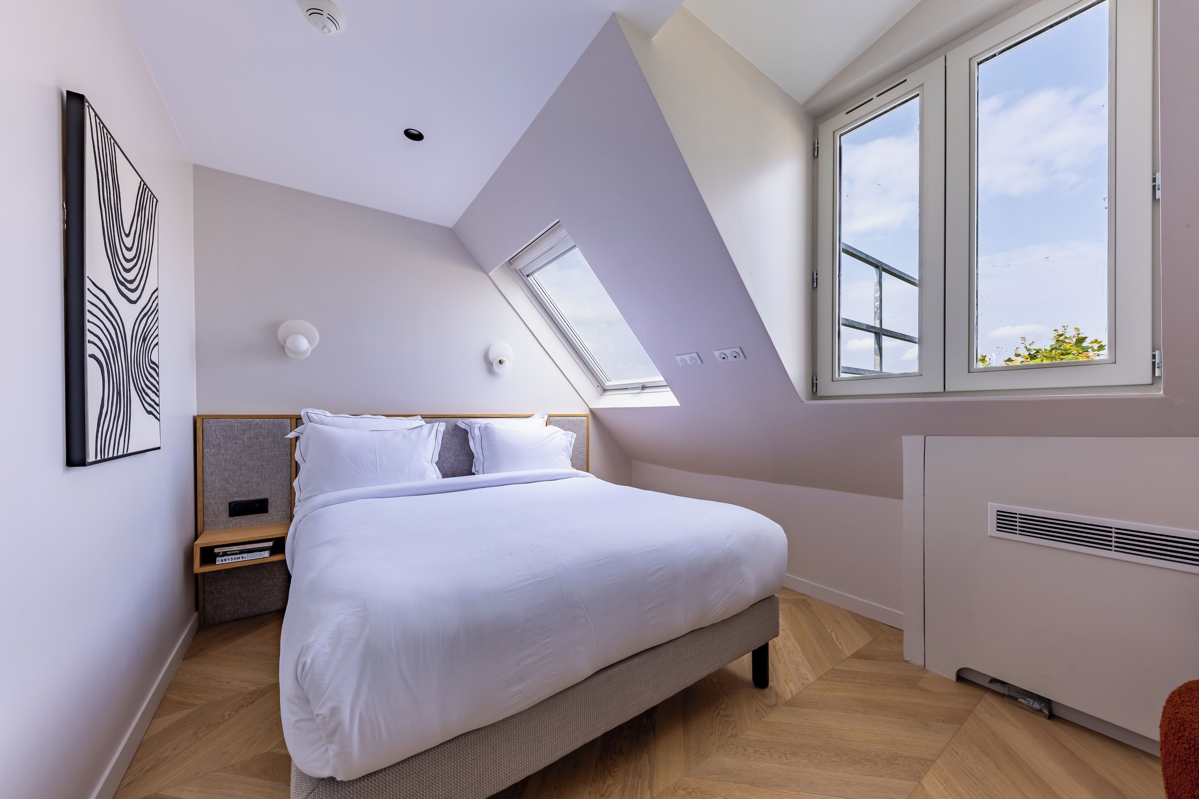 A modern bedroom with a sloped ceiling, a large bed, a nightstand, a wall-mounted light, and a window with a view of the sky.
