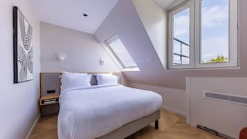 A modern bedroom with a sloped ceiling, a large bed, a nightstand, a wall-mounted light, and a window with a view of the sky.