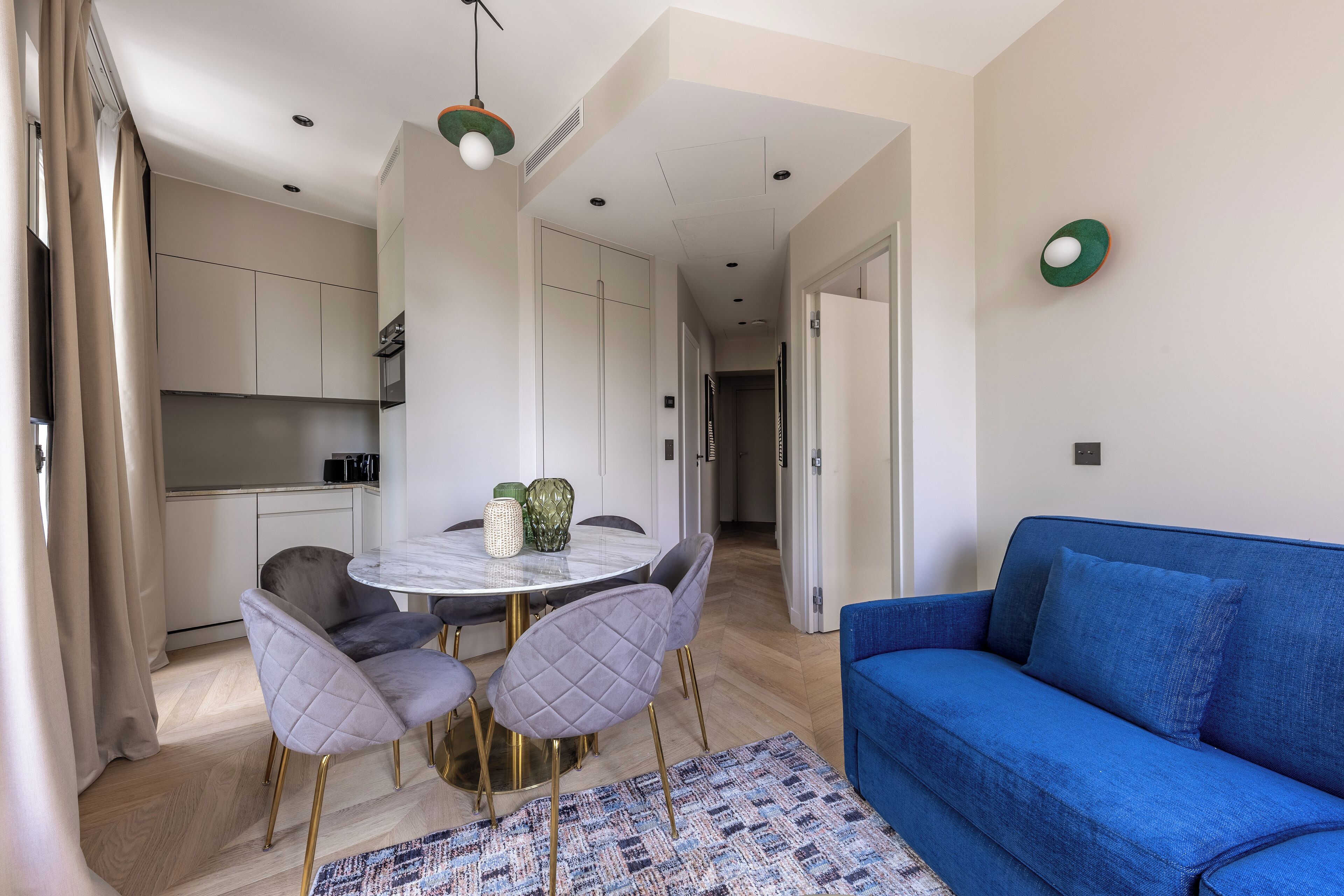 Un salon moderne avec un canapé bleu, une table de salle à manger ronde entourée de chaises, et un coin cuisine à l’arrière-plan.