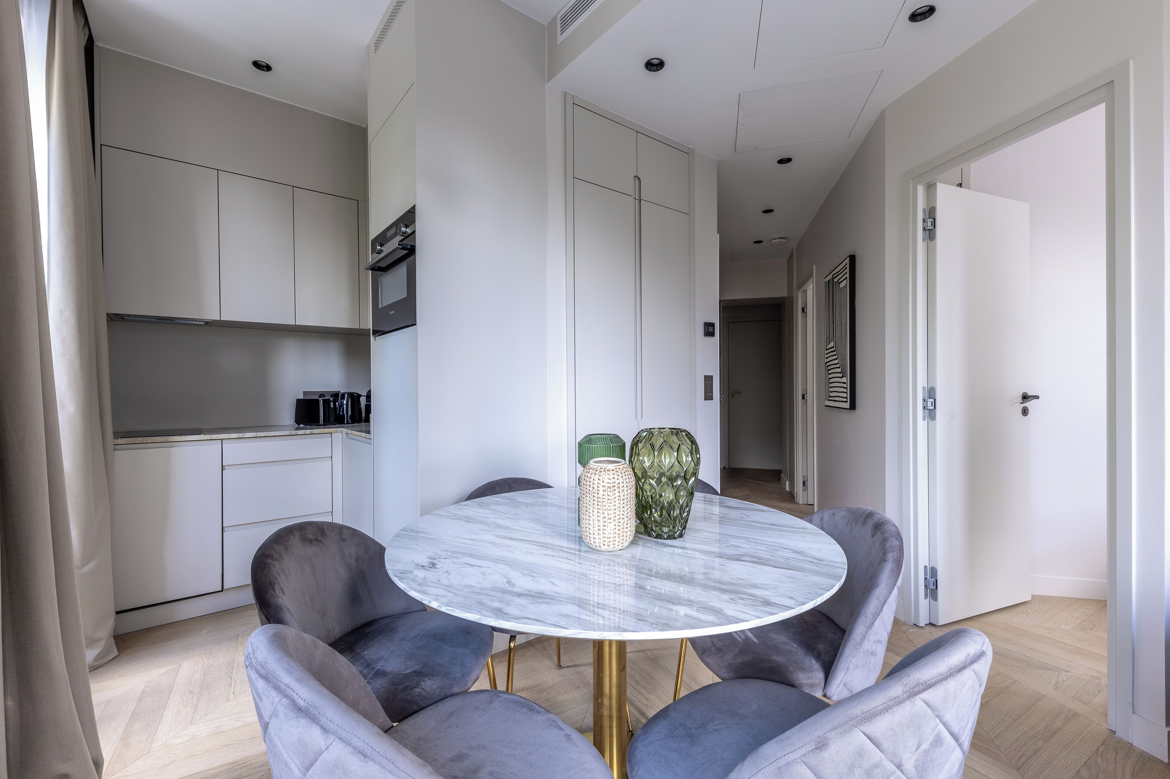 Una cocina moderna con armarios blancos, una zona de comedor con una mesa de mármol y sillas tapizadas en gris.