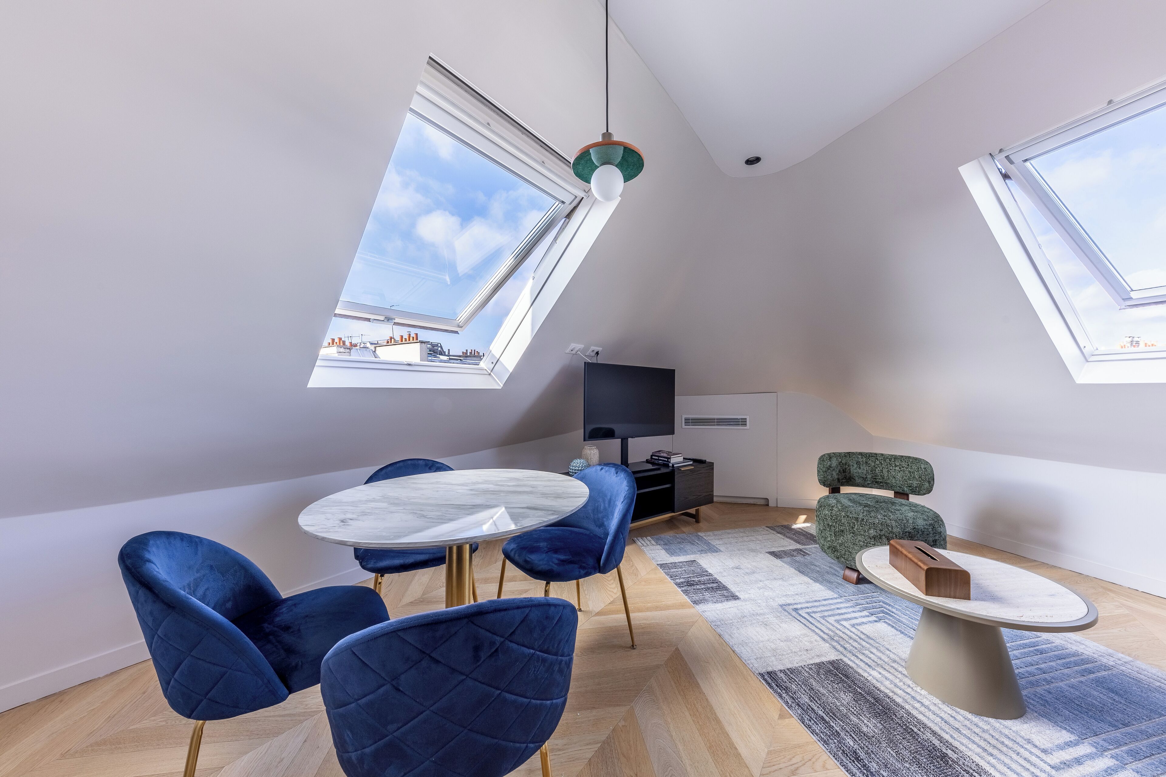 A modern loft bedroom with skylights, a round dining table, blue velvet chairs, a green upholstered chair, and a small side table.