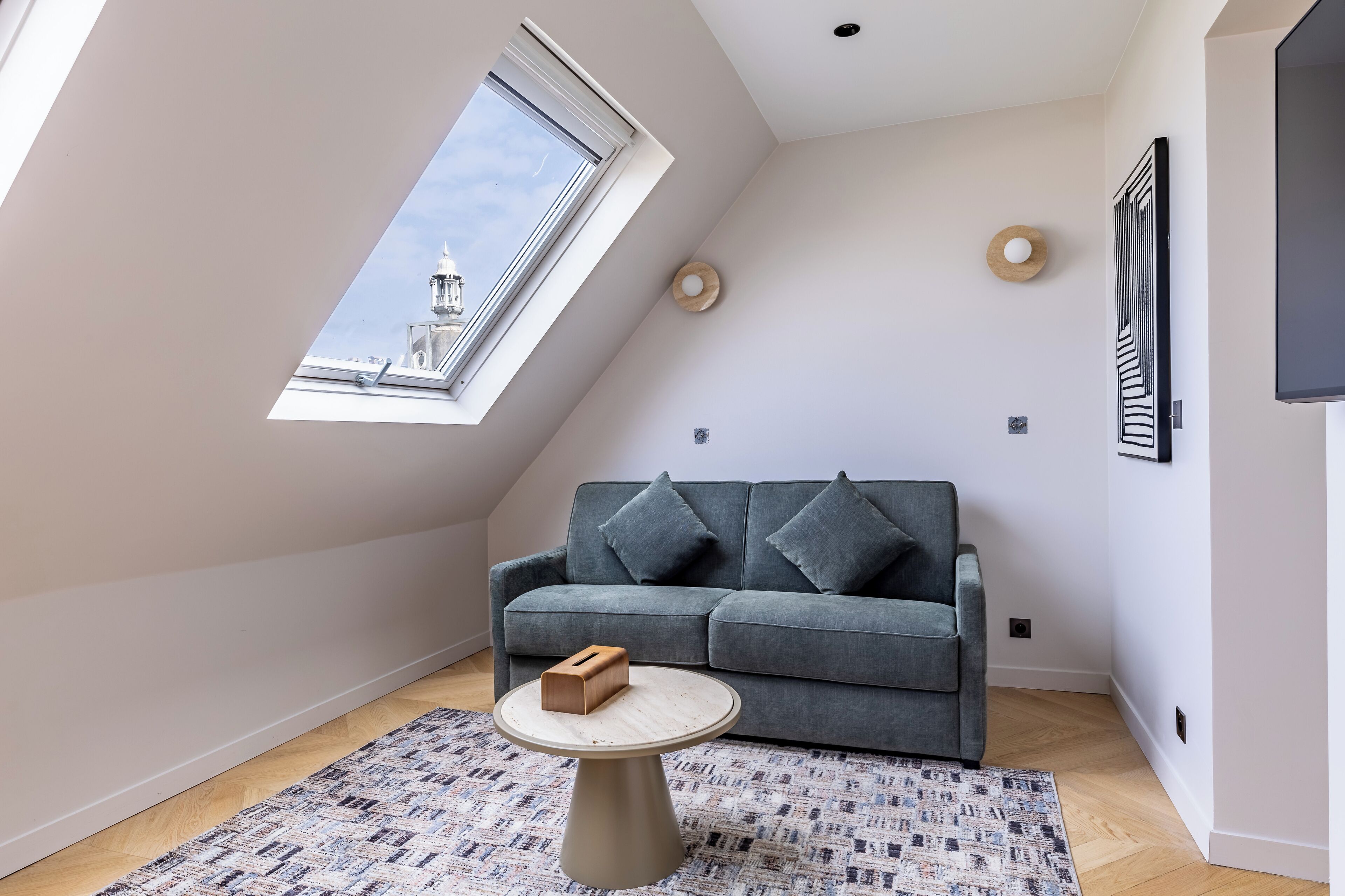 A modern loft apartment with a sloped ceiling, a grey sofa, a round coffee table, and a patterned rug.