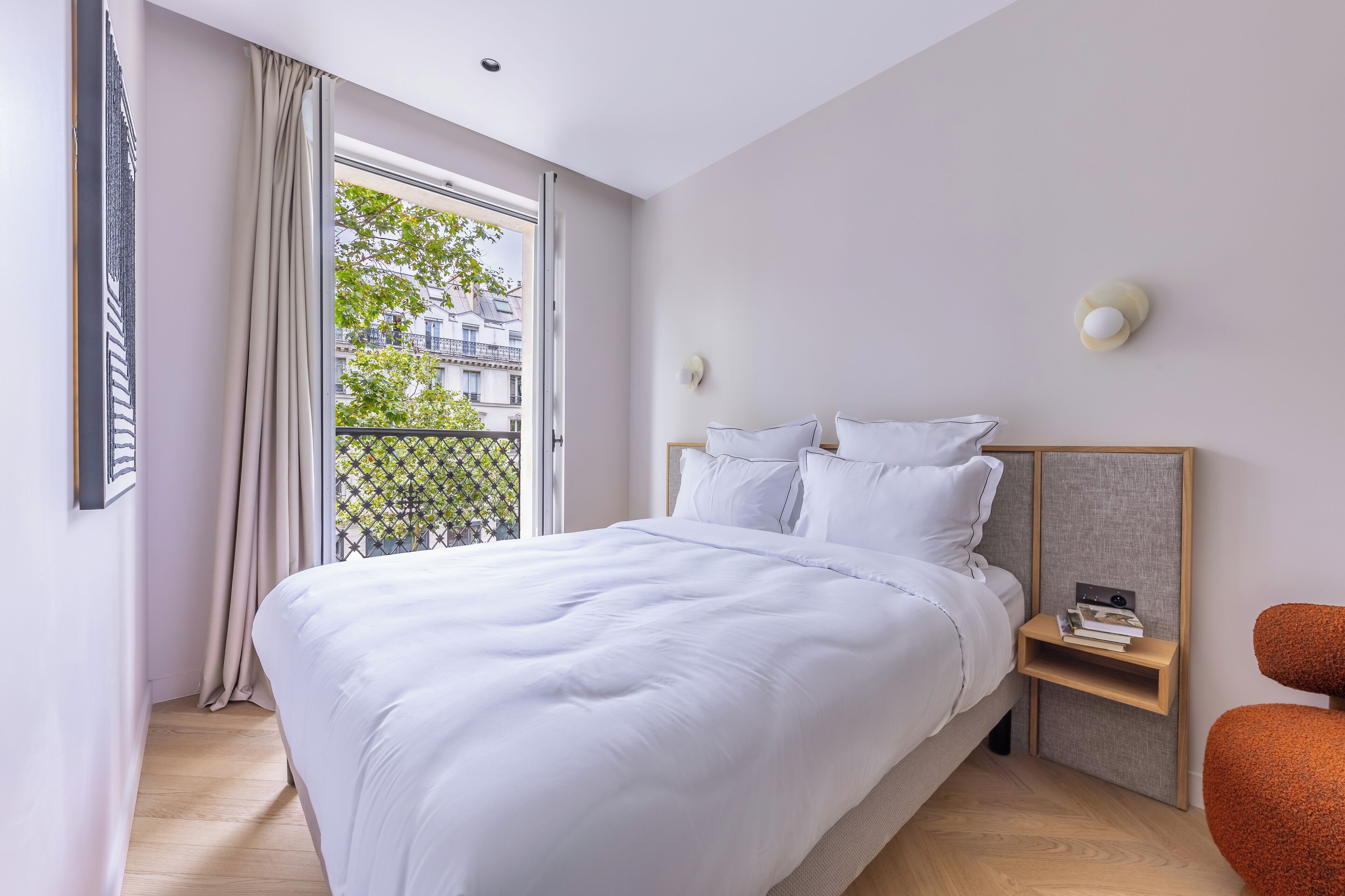 Een moderne slaapkamer met een groot bed, een nachtkastje met boeken en uitzicht op de stad door het raam.