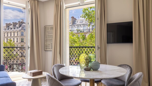 A modern living room with a round table, grey upholstered chairs, a flat-screen TV mounted on the wall, and a view of Parisian buildings through large windows. - Aparthotel Quartier Libre Marais (Paris)