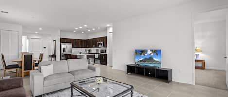 A modern living room with a sofa, coffee table, and TV. There is a kitchen area with dark wooden cabinets and a dining area with a table and chairs.