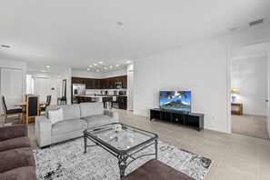 A modern living room with a sofa, coffee table, and TV. There is a kitchen area with dark wooden cabinets and a dining area with a table and chairs.
