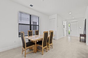A dining area with a wooden table and chairs, white walls, and a window with blinds.