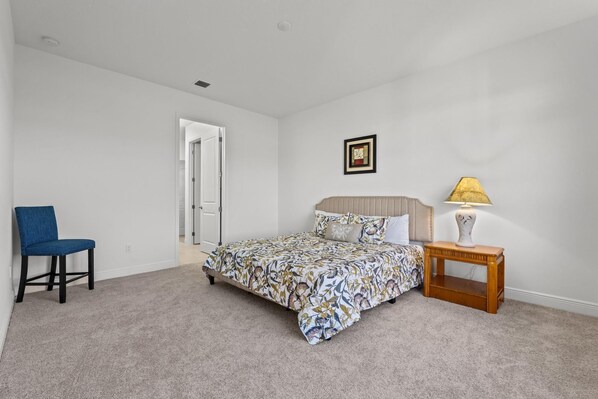 A bedroom with a bed, a chair, a bedside table with a lamp, and a door leading to another room.