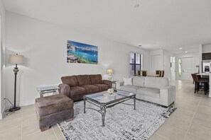A spacious living room with a brown sofa, a white sofa, and a glass coffee table. There is a painting of a coastal scene on the wall, and a dining area is visible in the background.