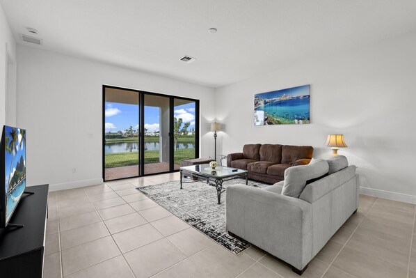 A modern living room with a large sliding glass door leading to a balcony, a flat-screen TV, a brown leather sofa, a grey sofa, a coffee table, and a painting of a coastal scene.