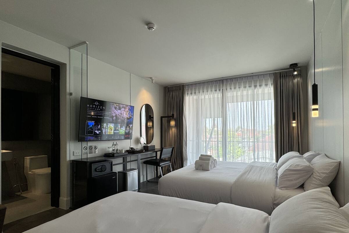 A hotel room with two beds, a desk with a TV, a mirror, and a view of the outside.