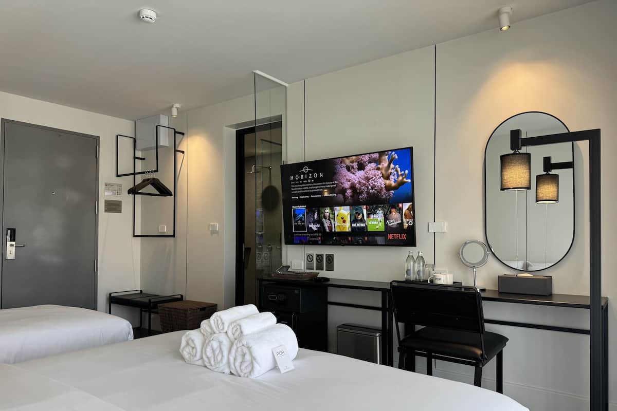 A hotel room with two beds, a desk with a flat-screen TV, a mirror, and a small seating area.