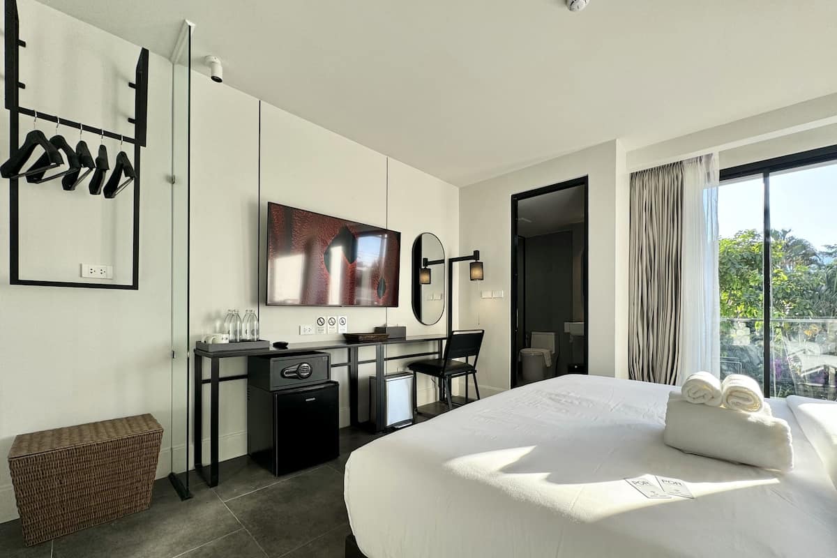 A modern hotel room with a large bed, a desk with a computer, a flat-screen TV, and a view of greenery outside.