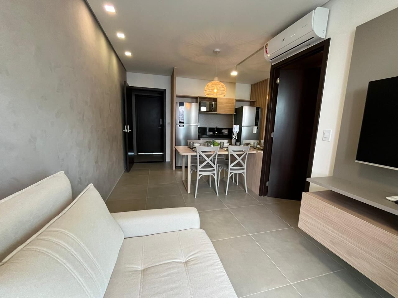 A modern living room with a sofa, a dining area with chairs, a kitchen with appliances, and a TV mounted on the wall.