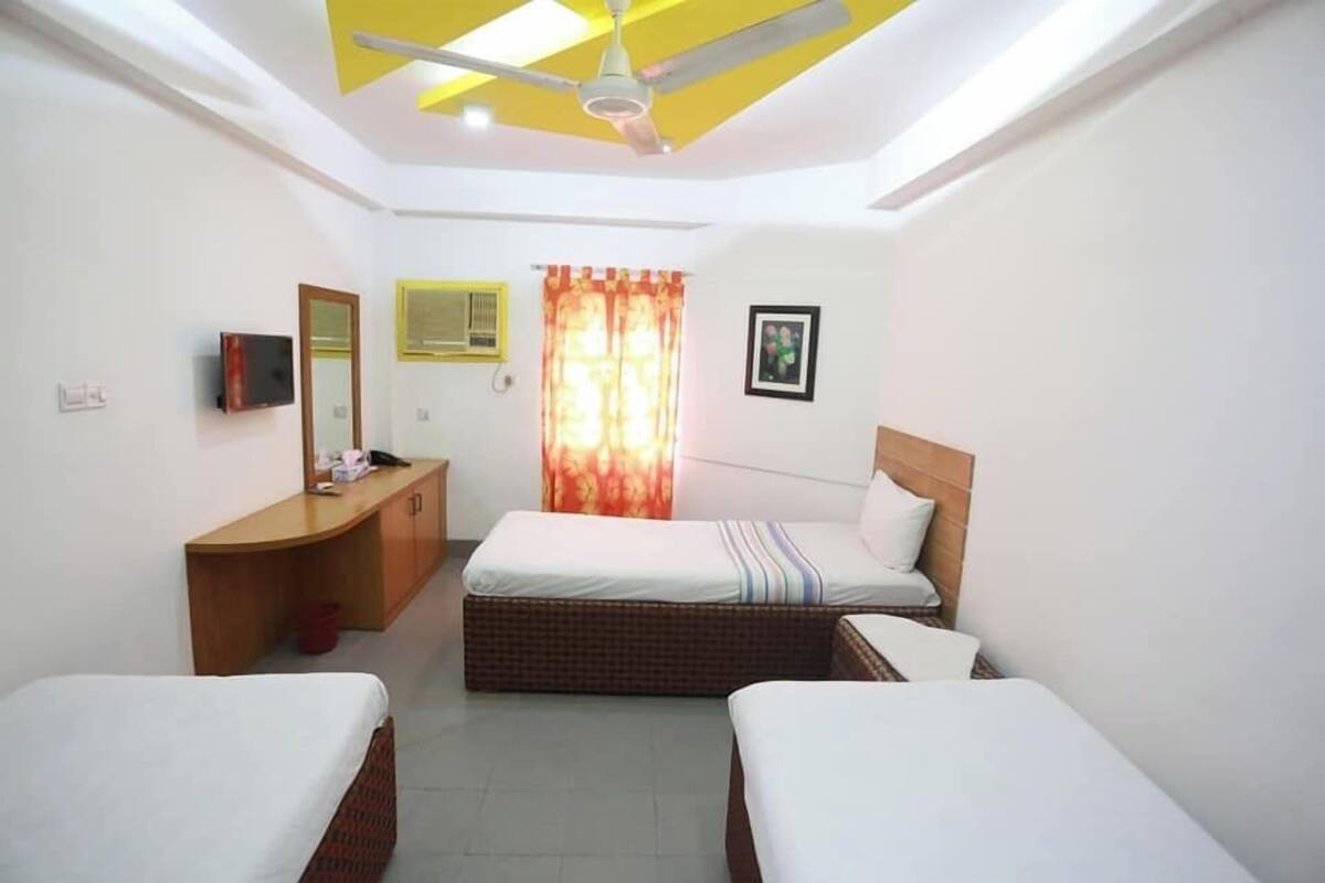 A hotel room with two single beds, a desk with a chair, a ceiling fan, a window with curtains, and a mounted TV.