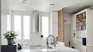A modern kitchen with a central island, white bar stools, a built-in bookshelf, and a large window. - Charmant Appartement en Plein Coeur des Batignolles (Paris)