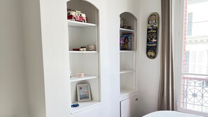 A bedroom with a bed, a dresser, built-in shelving with toys and books, and a window with a view of buildings. - Charmant Appartement en Plein Coeur des Batignolles (Paris)
