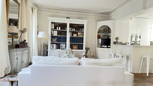 A living room with a white sofa, a bookshelf, a dining area, and a kitchen. - Charmant Appartement en Plein Coeur des Batignolles (Paris)