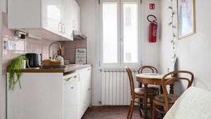 Kompakti keittiö, jossa on valkoiset kaapit, ruokapöytä tuoleineen, ikkuna sälekaihtimilla, palosammutin ja seinällä oleva kehystetty kuva.