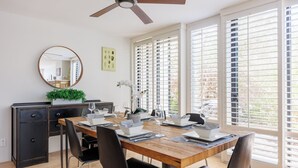 A dining area with a wooden table set for four, black chairs, a ceiling fan, and large windows with blinds. - Stunning Ocean View Townhome w/ Pool and Spa (Solana Beach)
