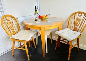 A round wooden dining table with two upholstered chairs, a bowl of fruit, a wine bottle, and a wine glass. - 2-bed Apartment in Coleshill (Birmingham)