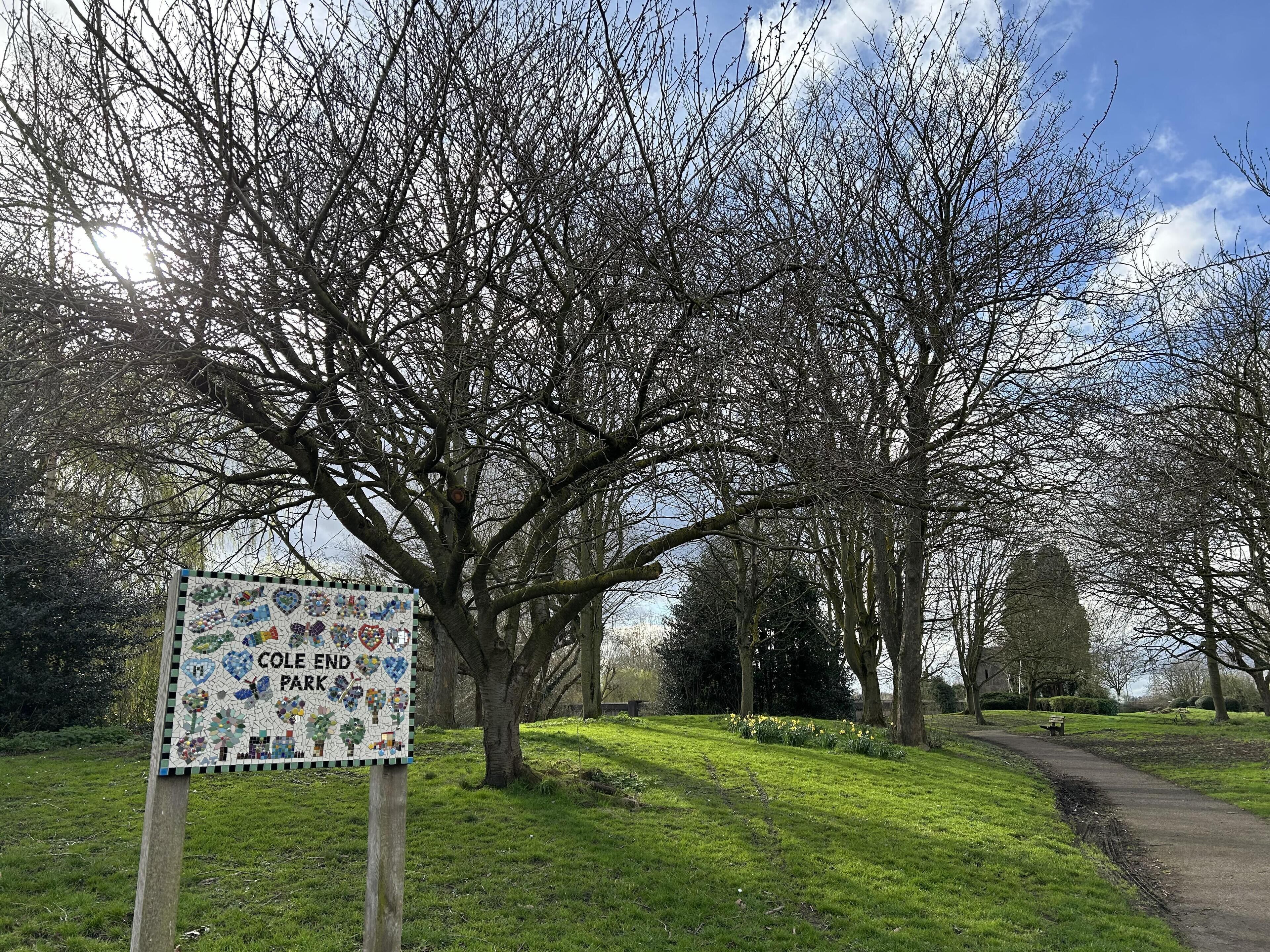 Un cartel de parque con diseño de mosaico, rodeado de árboles y un sendero.
