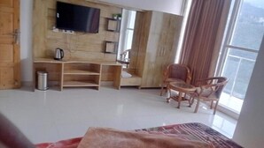 A living room with a TV mounted on a wooden wall, a small table with a chair, and a large window with curtains. - The Bel Mount (Dhanaulti)