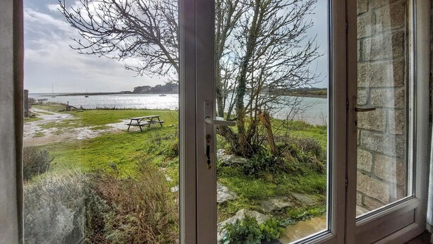 A view through a glass door to a grassy area with a picnic table, a stone wall, and a body of water with boats in the distance. - Gorgeous apartment in Locmariaquer with WiFi (Locmariaquer)
