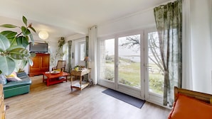 A spacious living room with a wooden floor, a sofa, a coffee table, and a television. There is a large window with curtains, and a view of the outdoors. - Gorgeous apartment in Locmariaquer with WiFi (Locmariaquer)